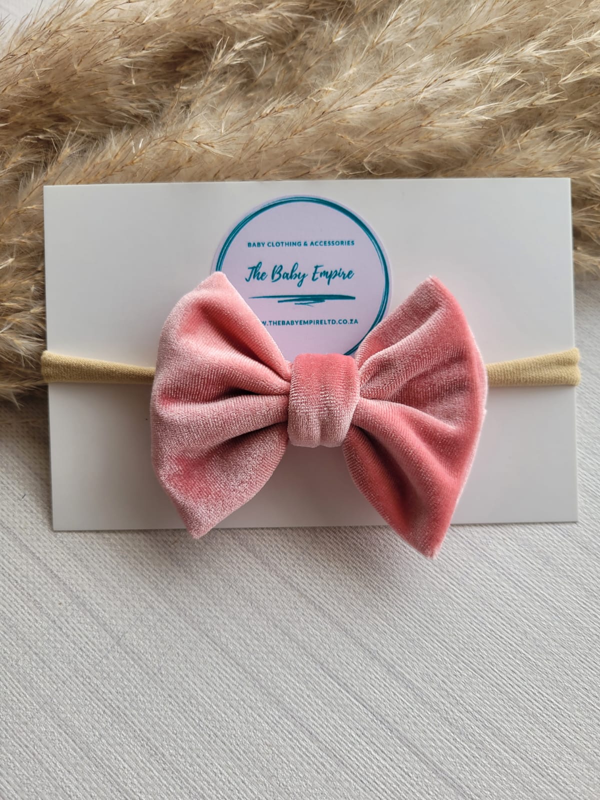 Pink velvet bow headband on a white card with 'The Baby Empire' logo, against a textured beige background.