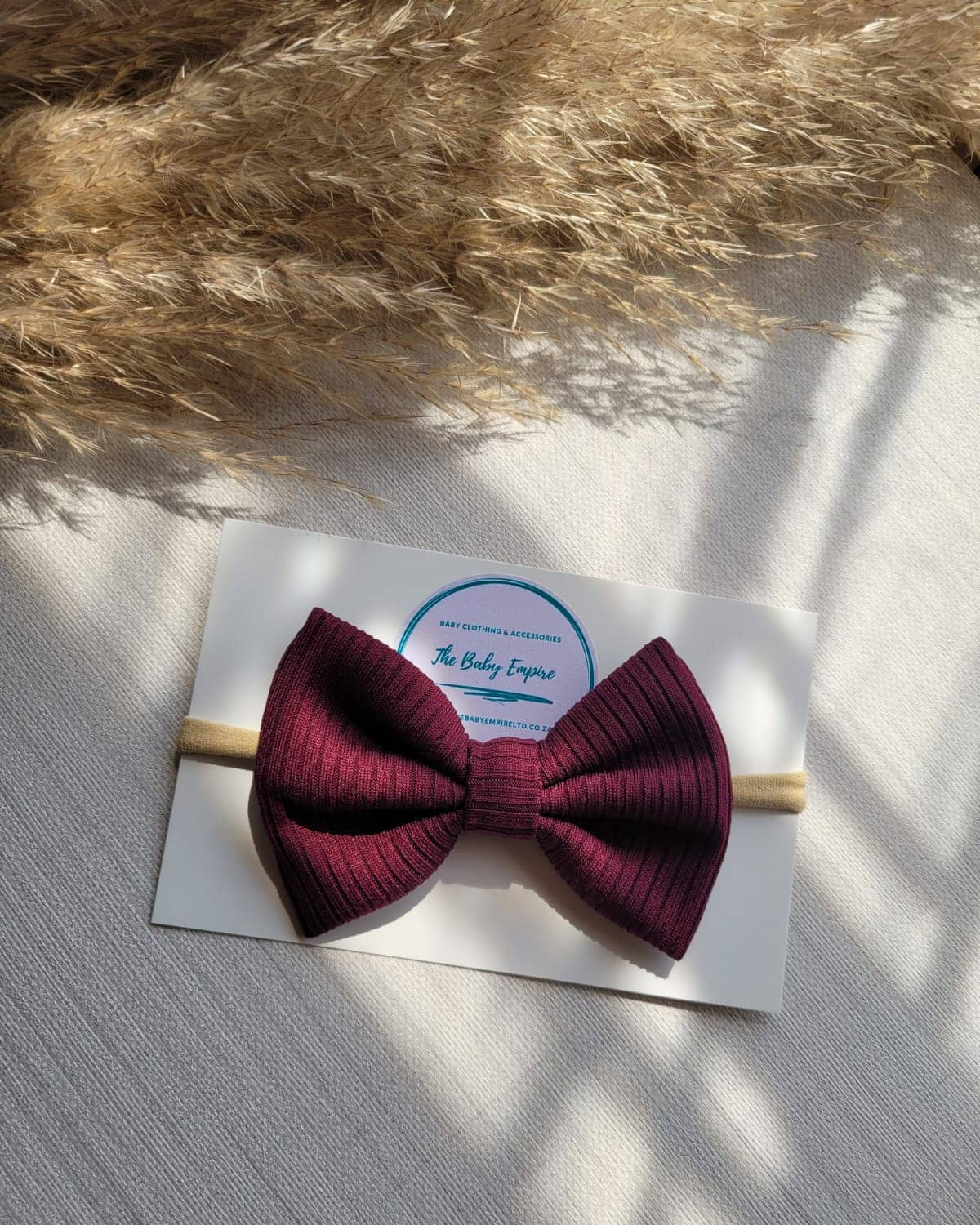 Maroon bow headband on a white card with a brand logo, placed on a textured surface with pampas grass.