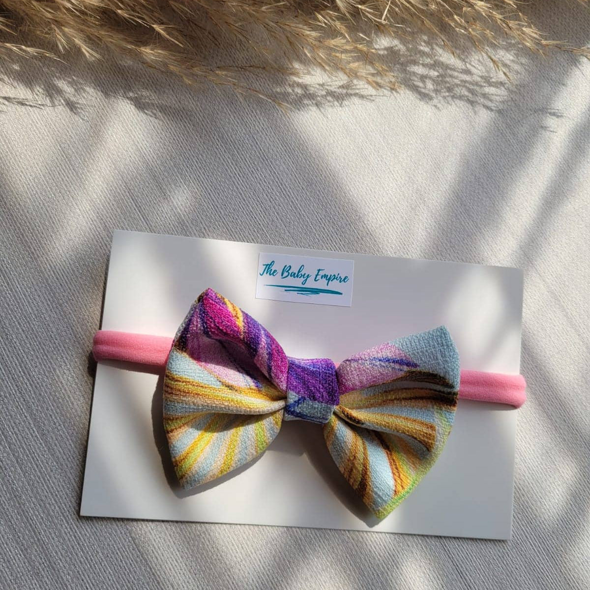 Colorful bow on a pink headband with  branding, placed on a textured surface.