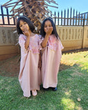 Two young girls in matching abayas standing outdoors with a wooden structure and fence in the background.