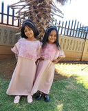 Two young girls in matching abayas standing outdoors with a wooden structure in the background.