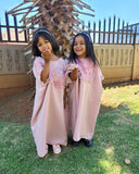 Two young girls in pink abayas standing outdoors with a decorative tree in the background.