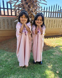 Two young girls in matching pink abayas standing outdoors with a decorative fence and tree in the background.