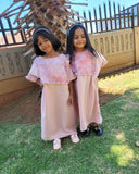 Two young girls in matching outfits standing on grass with a wooden structure in the background.