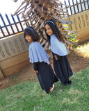 Two young girls in matching abayas standing outdoors with a wooden structure in the background.
