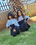 Two children in traditional attire sitting on grass with a wooden structure in the background.