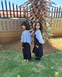 Two young girls in matching outfits standing in a grassy area with a wooden structure in the background.
