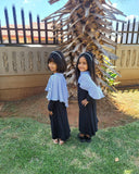 Two young girls in matching abayas standing in a grassy area with a wooden structure in the background.