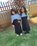 Two young girls in matching abayas standing on grass with a decorative wooden structure in the background.