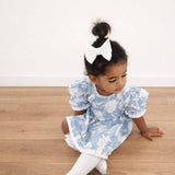 Young child wearing a blue floral dress with white puffed sleeves on a wooden floor.