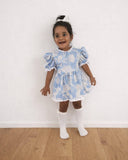 Child wearing a blue floral dress standing on a wooden floor with a white wall background
