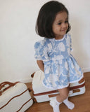Child in a blue floral dress standing next to vintage suitcases on a wooden floor.