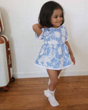 Child wearing a blue floral dress standing on a wooden floor with a white wall background
