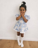 Young girl in a blue floral dress standing on a wooden floor against a white wall.