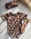 Leopard print baby onesie with matching headband and socks on a light wooden surface.