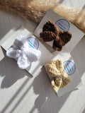 Three knitted hair bows in white, brown, and beige on a textured surface with labels.