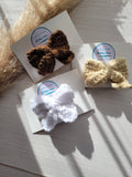 Three crocheted hair bows in brown, beige, and white on a textured surface with a brand label.