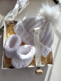 White baby hat, booties, and clip set in a decorative box on a light wooden surface.