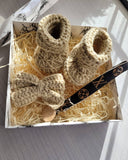 Pair of beige knitted baby booties with a bow in a box on a marble surface.