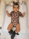 Baby wearing a leopard print outfit with a headband, lying on a white surface.