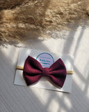 Maroon bow headband on a white card with a brand logo, placed on a textured surface with pampas grass.