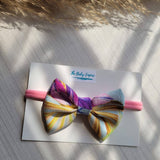 Colorful bow on a pink headband with  branding, placed on a textured surface.