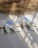 Two white bow hairbands on white cards with a brand label, placed on a textured surface.
